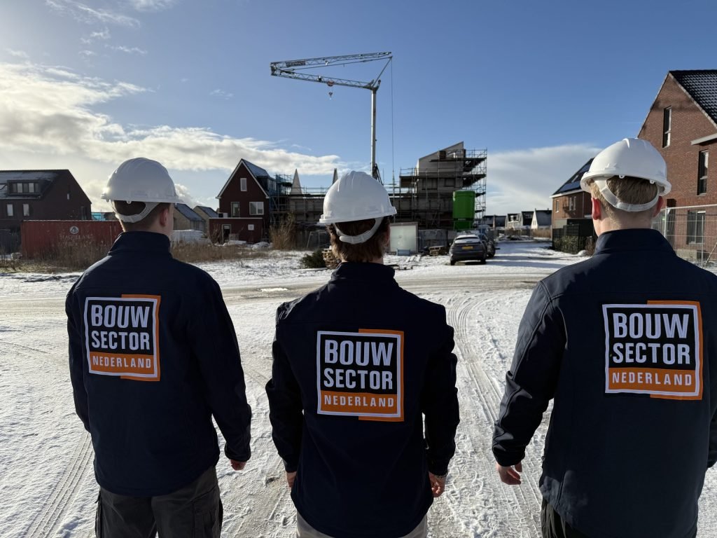 Drie vakmensen met witte helm en bedrijfsjas van Bouwsector Nederland, van achteren gezien, voor een bouwkraan